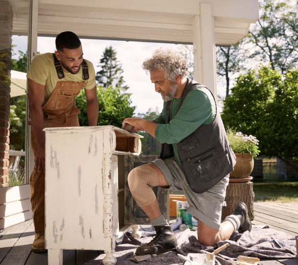 Man on bended knee wearing a knee support sands a piece of furniture while a younger man watches him. Man on bended knee wearing a knee support sands a piece of furniture while a younger man watches him.